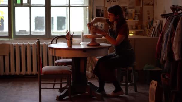 Ремесленница в уютной мастерской керамики, производящая гончарные изделия на рабочем месте со столом, колесом и инструментами — стоковое видео