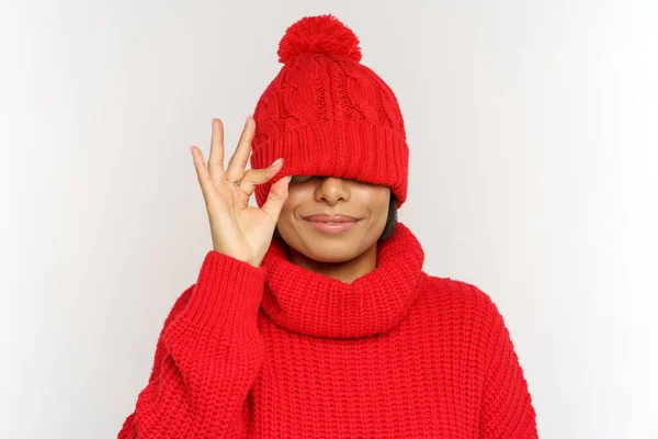 Brincalhão mulher africana engraçado se diverte vestindo chapéu de inverno e suéter cobrir os olhos sob tampa, tiro estúdio — Fotografia de Stock