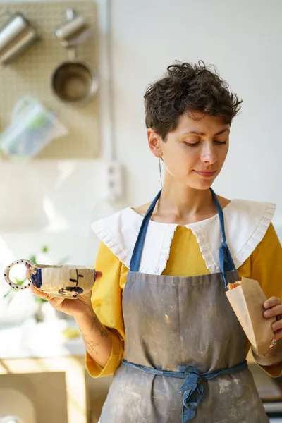 Zadowolona młoda kobieta ceramist pić kawę z ręcznie robionej filiżanki garncarskiej i ma przekąskę w pracowni garncarskiej — Zdjęcie stockowe