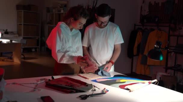 若いデザイナーの男性と女性のドレスメーカーは一緒にスタジオワークショップで新しい服コレクションに取り組んでいます — ストック動画