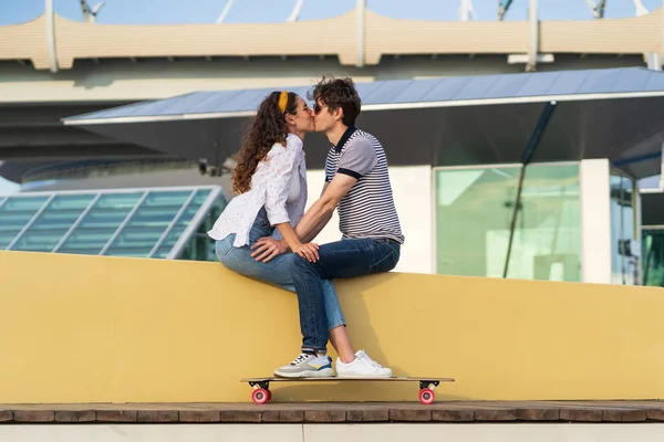 Стильная пара целуется, сидя в скейтпарке. Молодые симпатичные лонгбордисты мужчина и женщина в любви обнимаются — стоковое фото