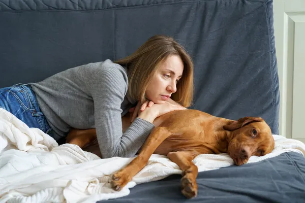 Una donna che si prende cura di un vecchio cane malato. Donna infelice sdraiato sul divano abbracciando stanco vizsla malato dopo la malattia — Foto Stock