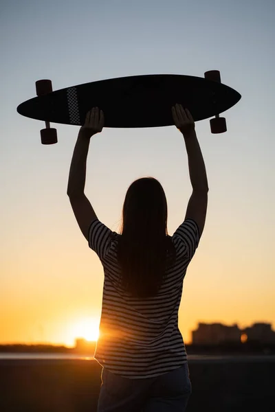 Ελευθερία και αστικός τρόπος ζωής: έφηβος κορίτσι σκέιτερ με longboard ματιά στο ηλιοβασίλεμα πίσω όψη — Φωτογραφία Αρχείου