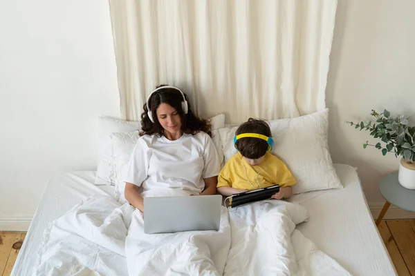 Mom typing on laptop computer lying in bed with preschool son kid watch video or cartoon on tablet — Stock Photo, Image