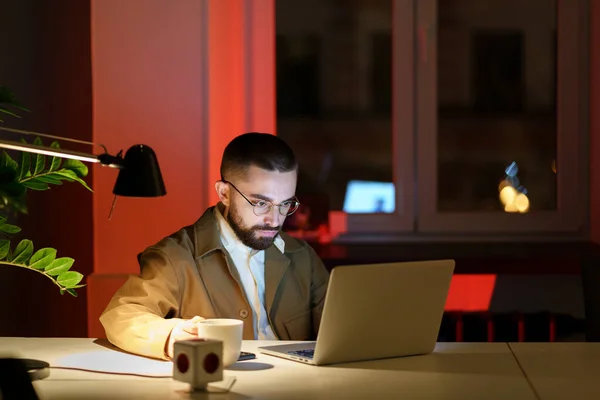 Soustředěný muž se dívá na laptop pít kávu na pracovišti v kanceláři za tmy pozdě v noci přepracování — Stock fotografie
