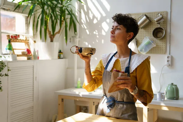 Joven artesana creativa disfrutando del desayuno mientras comienza a trabajar en su propio estudio de cerámica por la mañana —  Fotos de Stock