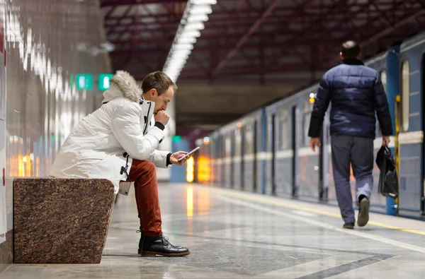 電話を見てうつ病の若い男を泣かせることは 悪い知らせを受け メッセージを受け取り 手で口を覆い 地下鉄の駅のベンチに座って 背景に乗客 関係の問題 — ストック写真