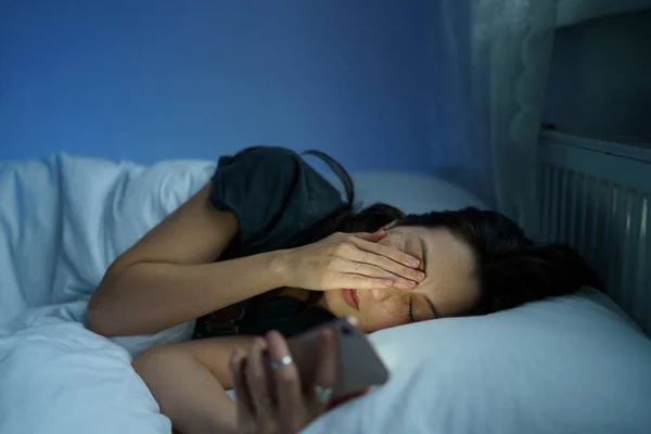 Vyčerpaná žena ležící v posteli bezesné držení smartphone číst o poruchách spánku a duševní zdraví — Stock fotografie