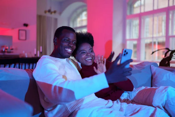 Cheerful african couple talk by video call on smartphone with friends while relaxing on sofa at home