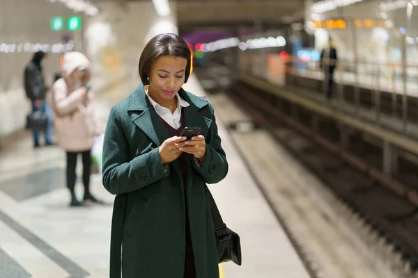 Siyahi iş kadını tren istasyonunda, akıllı telefondan mesaj ya da iş e-postası okumak için bekliyor. — Stok fotoğraf
