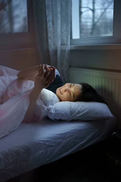 Влюбленная молодая женщина переписывается с парнем из приложения для свиданий всю ночь напролёт с мечтательной улыбкой в постели — стоковое фото