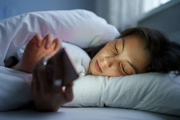 Молодая женщина, лежащая ночью в постели с мобильным телефоном, страдает бессонницей после расставания — стоковое фото