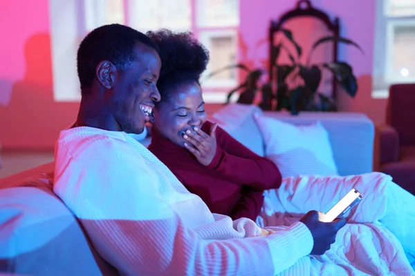 Feliz pareja de la familia africana riendo mientras se relaja en el sofá y mirando la pantalla del teléfono inteligente —  Fotos de Stock