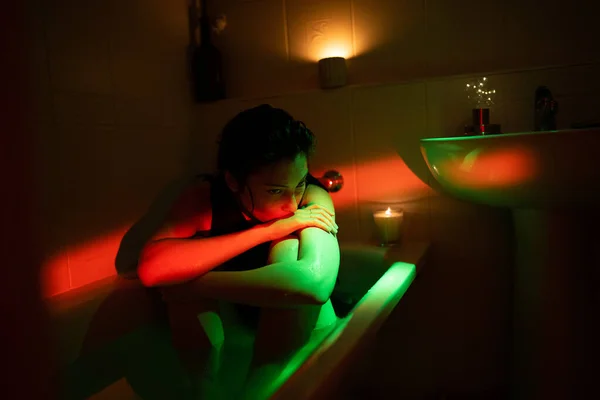 Young depressed lonely woman sitting in bathtub under neon light, suffering from breakup depression