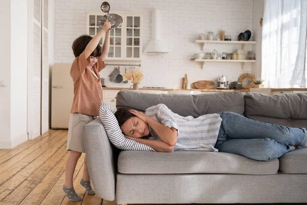 Agotada madre soltera sufren de dolor acostado en el sofá con travieso hijo ruidoso golpeando utensilios de cocina — Foto de Stock