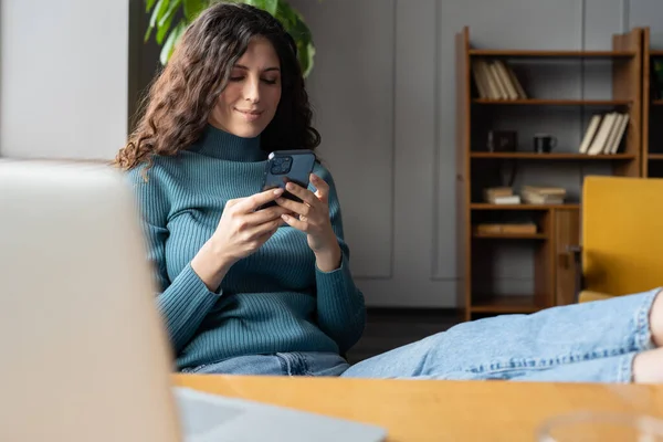 Mladá usměvavá žena drží smartphone chatování on-line při relaxaci v útulné domácí kanceláři — Stock fotografie