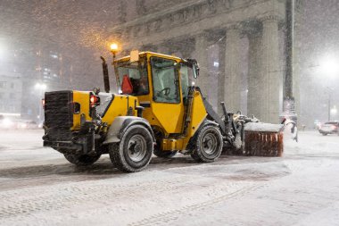 Kış servis araçları. Büyük sarı kar küreme aracı, gece ağır kar yağışı altında şehirde çalışıyor.