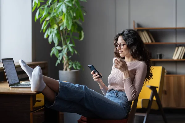 Uvolněná podnikatelka pije kávu, čte online zprávy na smartphonu při relaxaci na pracovišti — Stock fotografie