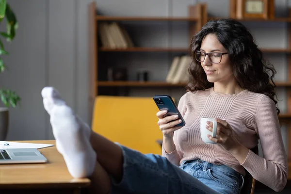 Νεαρή χαλαρή γυναίκα υπάλληλος απολαμβάνει διάλειμμα στη δουλειά, χρησιμοποιώντας smartphone και πίνοντας τσάι στο χώρο εργασίας — Φωτογραφία Αρχείου