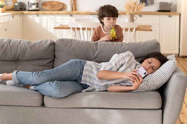 Calm young woman mother, nanny ignore noisy child lying on sofa using smartphone chatting in apps — ストック写真