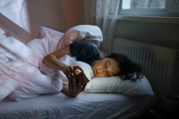 Young depressed woman in bed staring at smartphone screen, feeling envious on social media — Stock fotografie