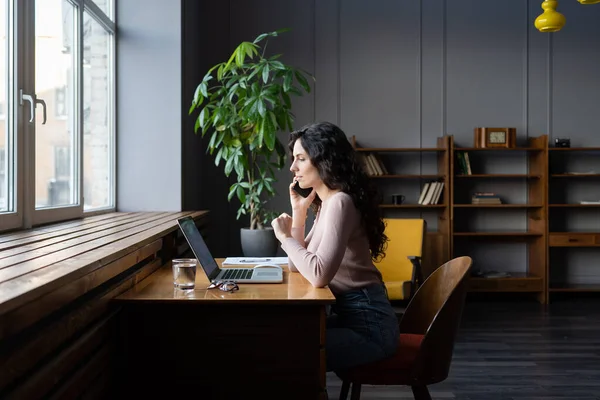 Ung affärskvinna arbetar på distans på bärbar dator i coworking utrymme, samtal med kunden via mobiltelefon — Stockfoto