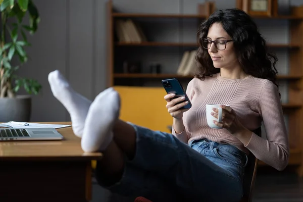 Ontspannen jonge vrouw freelancer rusten aan het bureau met mobiele telefoon en koffiekop, genieten van freelance — Stockfoto