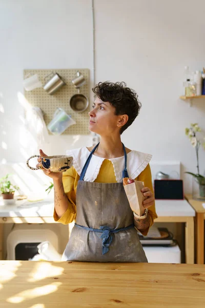 Przerwa na kawę w studio: kobieta garncarz pije herbatę z kubka rzemiosła w kreatywnym warsztacie ceramiki — Zdjęcie stockowe