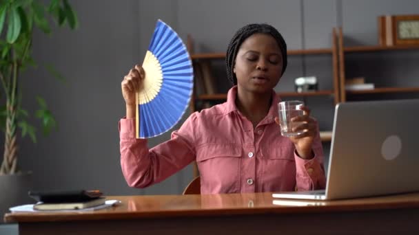 Overwhelmed black woman suffer from anxiety attack, dyspnea or heat at home drink water waving fan — Stock Video