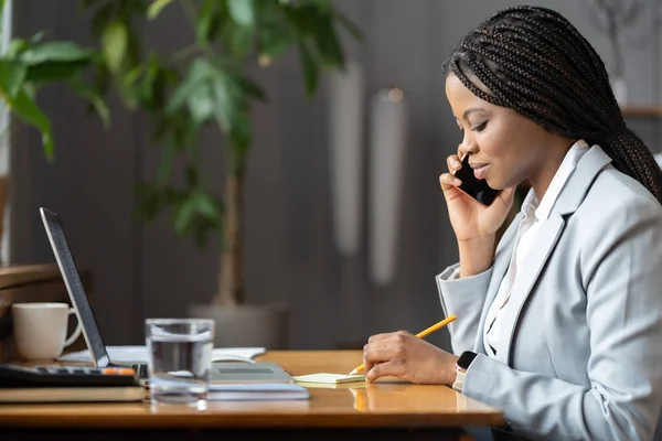 穿着西服的严肃的非洲女人在舒适的工作环境中一边打电话一边记笔记 — 图库照片