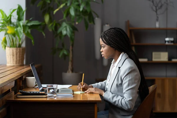 在笔记本电脑上工作的非洲裔女青年学员在办公室写字台为商业报告做笔记 — 图库照片