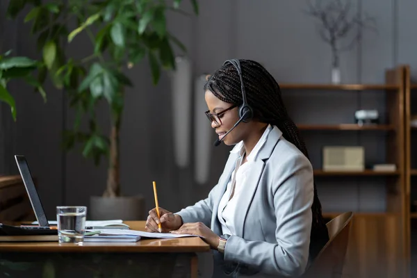 Joyeuse femme d'affaires noire dans l'étude casque en ligne écrire des notes regarder webinaire cours sur ordinateur portable — Photo