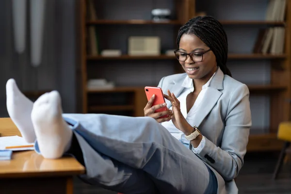 Heureuse femme africaine assise avec les jambes au bureau défilant à travers les médias sociaux sur smartphone — Photo