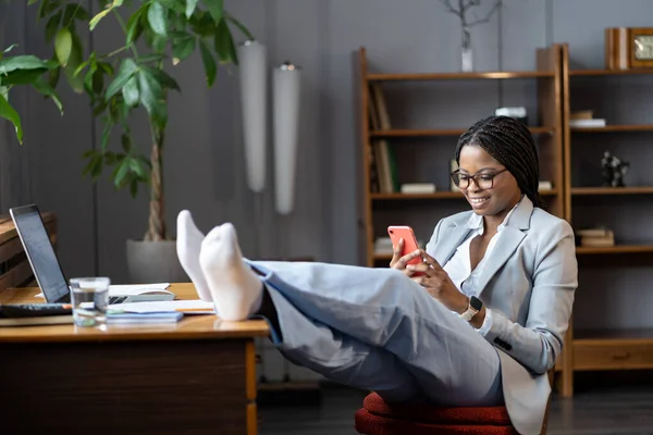 Afro détendue femme d'affaires procrastiner sur le lieu de travail s'asseoir avec les pieds sur le bureau utiliser la messagerie smartphone — Photo