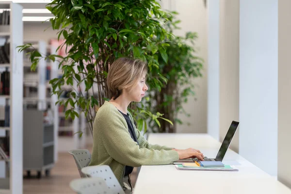 Professor universitário pensivo trabalhando no laptop na biblioteca vazia acolhedora cheia de plantas de sala verdes — Fotografia de Stock