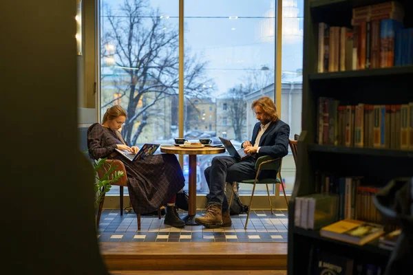 Mujer aburrida y hombre serio centrado sentados juntos en la mesa en la cafetería por la ventana de trabajo en los ordenadores portátiles — Foto de Stock