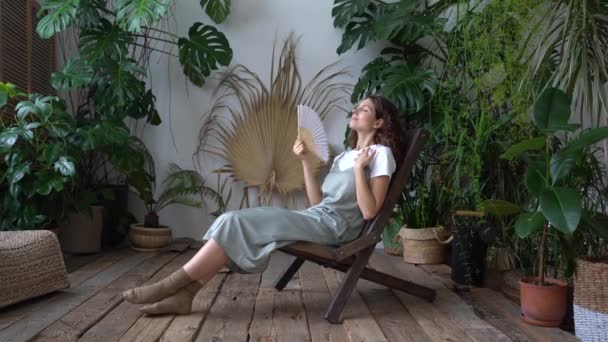Fleuriste femme heureuse rafraîchissant dans le jardin à la maison respirant l'air frais tenir ventilateur de papier à la main à la journée chaude — Video