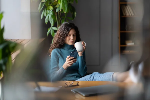 Glad kvinna förhalar på arbetsplatsen dricka kaffe läsa online nyheter på smartphone på kontoret skrivbord — Stockfoto