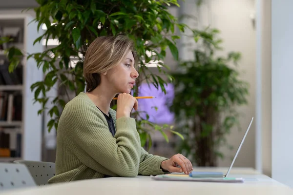 Professeur féminin d'âge moyen pensif préparant des documents de cours sur ordinateur portable à la bibliothèque de l'Université — Photo