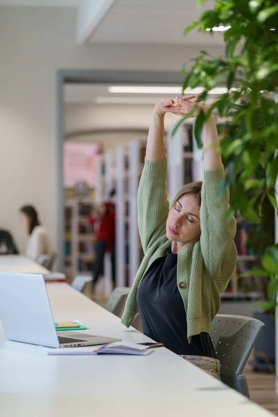 Femme fatiguée apprenant adulte étirant les bras les yeux fermés tout en étudiant dans une bibliothèque confortable — Photo