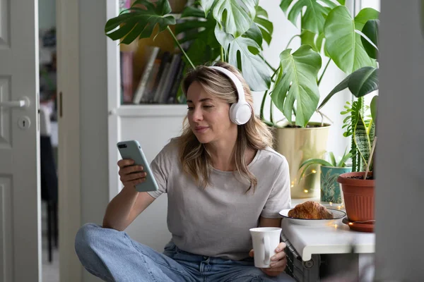 Kobieta w średnim wieku słuchająca muzyki w słuchawkach, korzystająca ze smartfona, rozmawiająca w mediach społecznościowych. — Zdjęcie stockowe