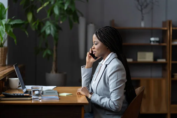非洲女商人打电话咨询客户，坐在办公桌前，办公室里有手提电脑 — 图库照片