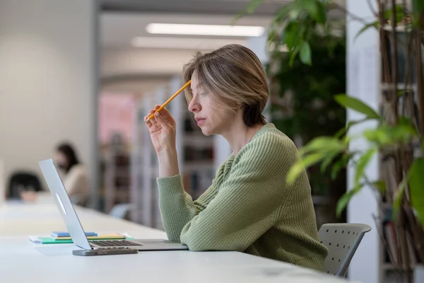 Концентрована студентка-зріла вивчає онлайн на ноутбуці в сучасній бібліотеці в оточенні зеленого — стокове фото