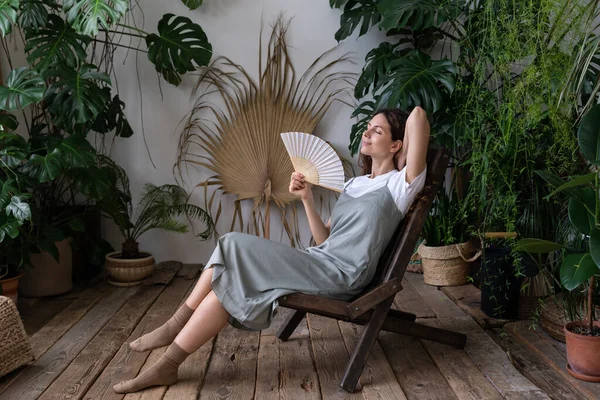 夏日炎热的日子里，在舒适的室内花园里，坐在扶手椅上静坐着的女人可以从风扇里呼吸新鲜空气 — 图库照片
