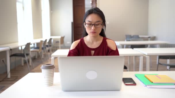 Focalisé Cher asiatique fille dans des lunettes étudier en ligne sur ordinateur portable tout en étant assis à la table de la bibliothèque — Video