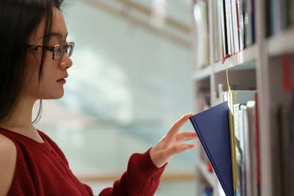 Азиатская студентка забирает книгу с полки, стоя между книжными шкафами. — стоковое фото