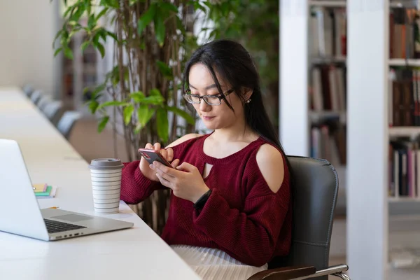Student flicka procrastinate i biblioteket med smartphone chatt i sociala medier inte förbereder sig för examen — Stockfoto