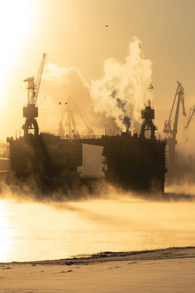 Grues du chantier naval en hiver givré, vapeur au-dessus de la rivière, surface lisse de la rivière au coucher du soleil. — Photo