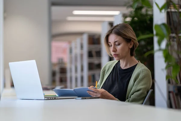 Концентрированная женщина-преподаватель университета проверяет расписание курсов, сидя в пустой библиотеке — стоковое фото