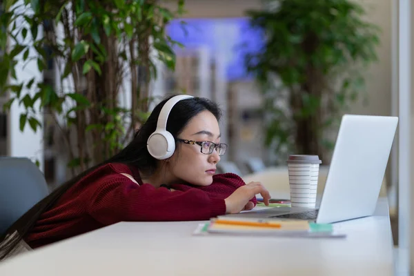 Cansado entediado asiático estudante menina em fones de ouvido surf internet no laptop enquanto estuda na biblioteca — Fotografia de Stock